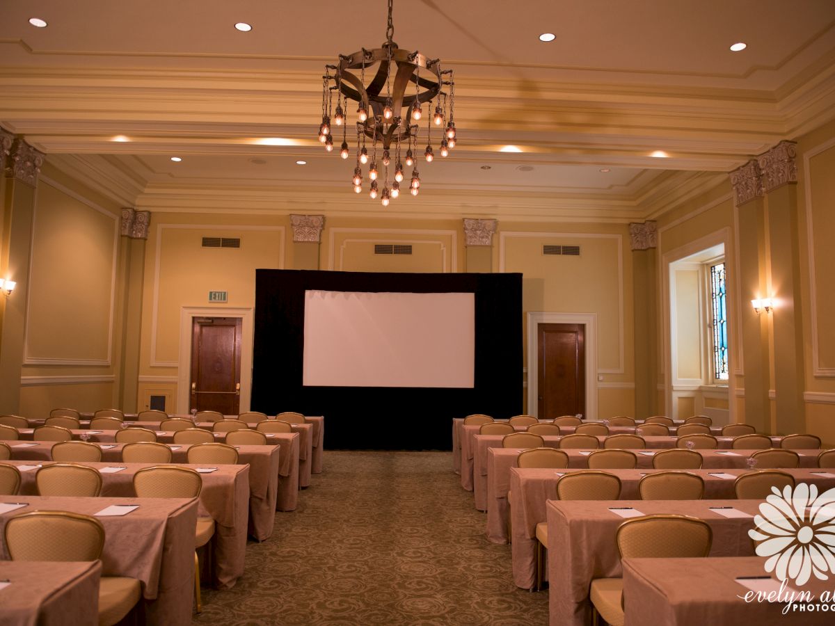 A conference room with rows of chairs and tables, facing a projector screen, decorated with a chandelier and soft lighting, ending the sentence.