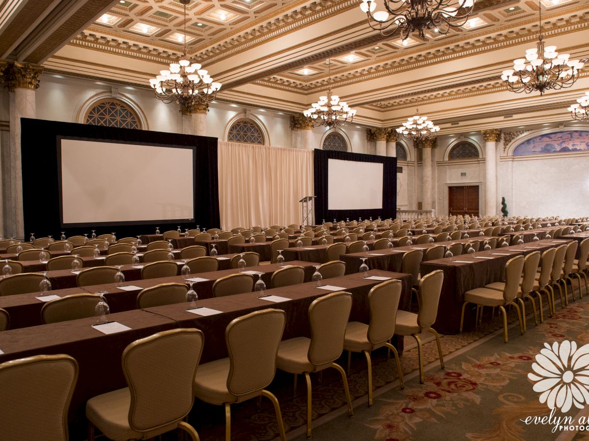 A large conference room with ornate chandeliers, two projection screens, rows of chairs, and tables set up with notepads and pens, ready for an event.