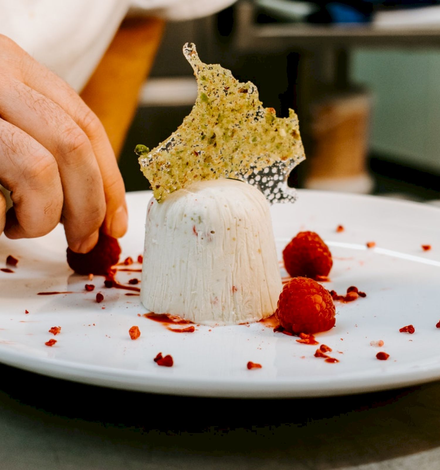 A hand is placing raspberries on a white dessert, garnished with a delicate, crispy decoration on a white plate.