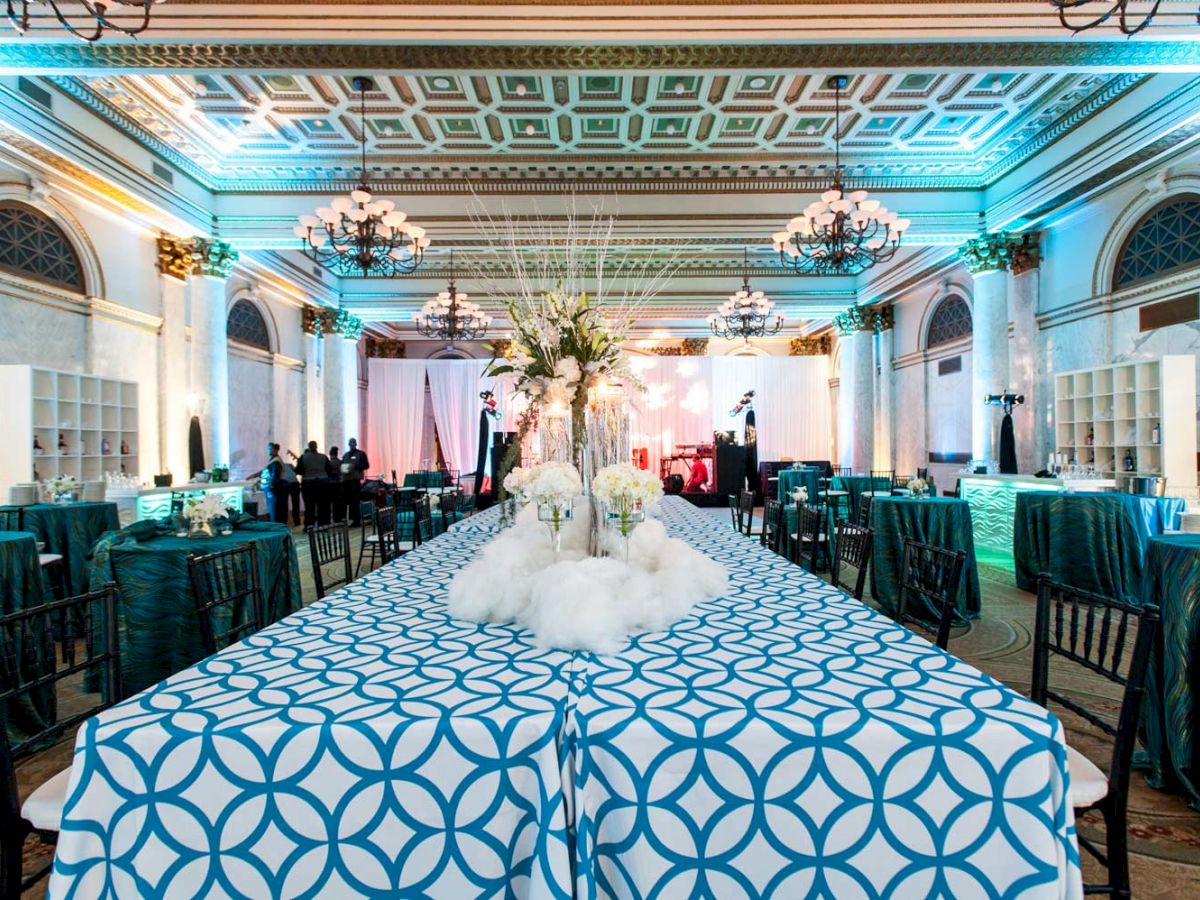 The image shows a large elegant hall set for an event, with long tables covered in geometric-patterned tablecloths, decorated with floral centerpieces.