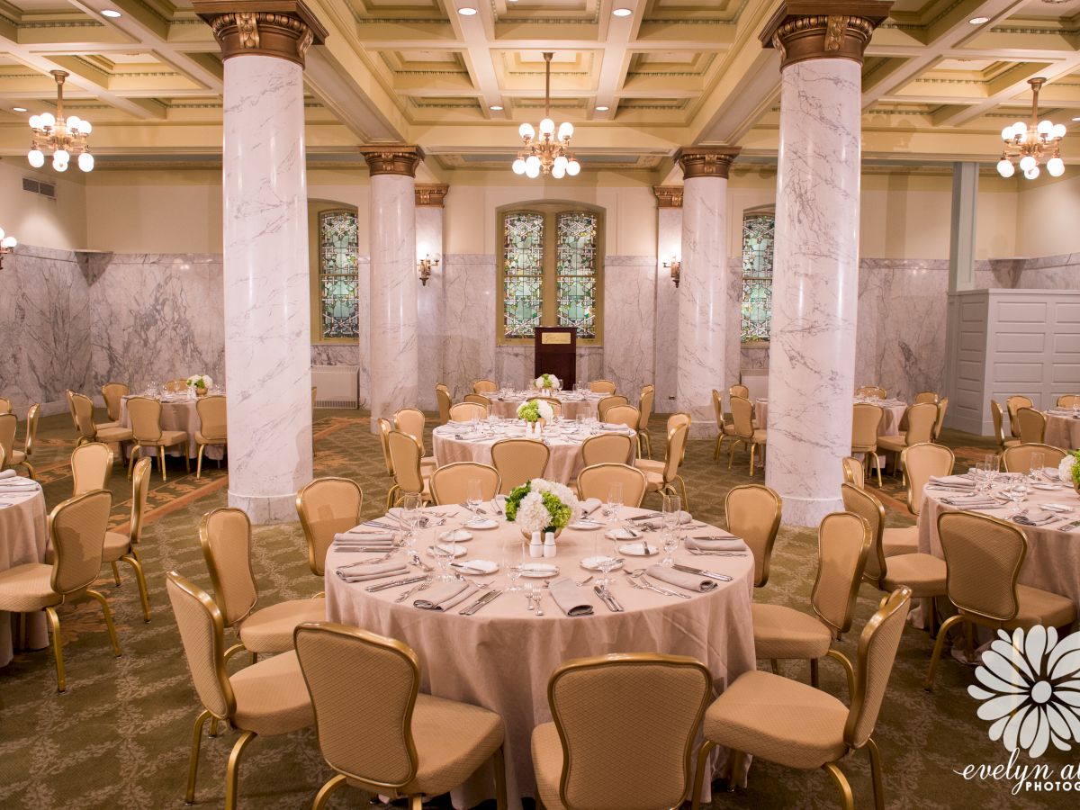 The image depicts an elegant banquet hall set with round tables covered in beige tablecloths, with floral centerpieces and surrounded by chairs.