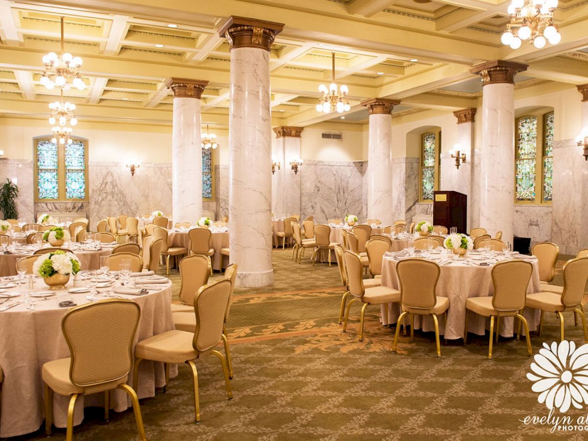 An elegant banquet hall with round tables, beige chairs, floral centerpieces, and chandeliers. Marble columns and decorated ceiling complete the setting.