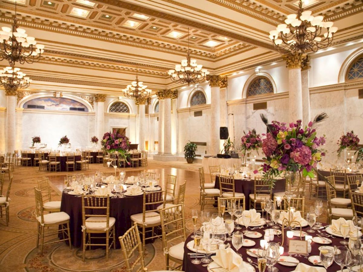 The image depicts an opulent banquet hall with elegantly decorated round tables, chandeliers, gold chairs, and floral centerpieces.