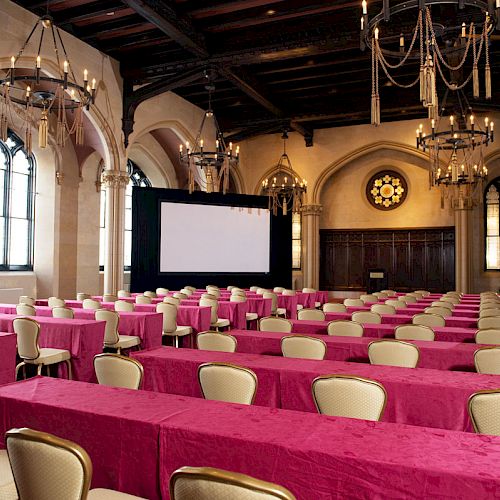 A conference room with pink tablecloths, cream chairs, chandeliers, stained-glass windows, and a large projector screen at the front.