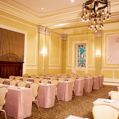 The image shows an elegant room set up for a meeting or event, with rows of tables and chairs, chandeliers, and a stained glass window.