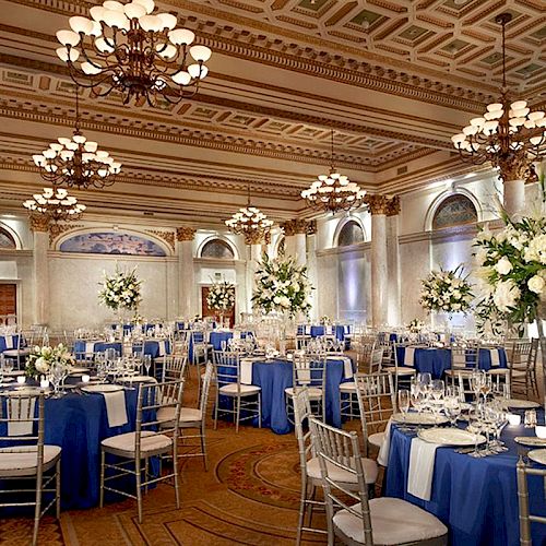 A lavishly decorated banquet hall with blue and white table settings, elegant chandeliers, and tall floral centerpieces.