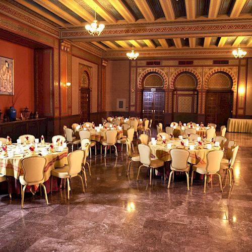 A banquet hall is set up with round tables and chairs, chandeliers, and intricate decor. Lighting creates a warm ambiance in the spacious room.