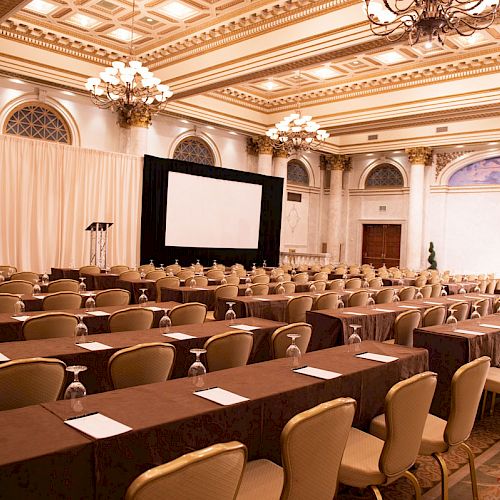 The image shows an elegant conference room with rows of tables and chairs, notepads, and water glasses, ready for an event or presentation.