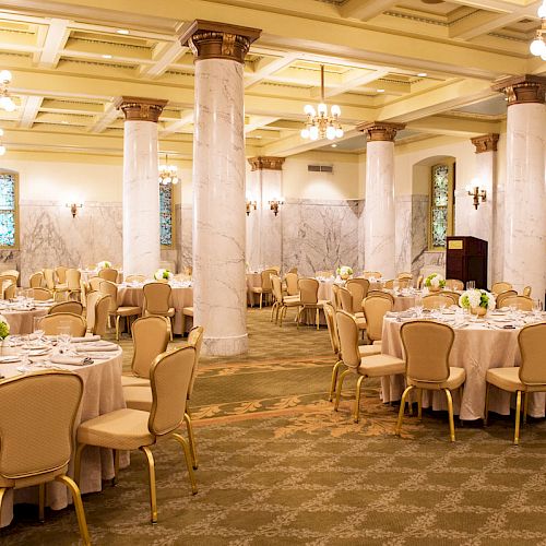 This image shows an elegantly decorated banquet hall with round tables, beige chairs, chandeliers, marble columns, and floral centerpieces on the tables.