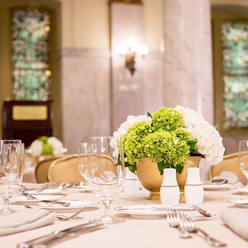 Elegant table setting with white linens, glassware, cutlery, and a centerpiece of white and green flowers in a gold vase.