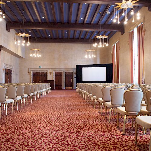 A large conference room with rows of white chairs facing a screen, decorated with chandeliers and curtains, features a patterned carpet.