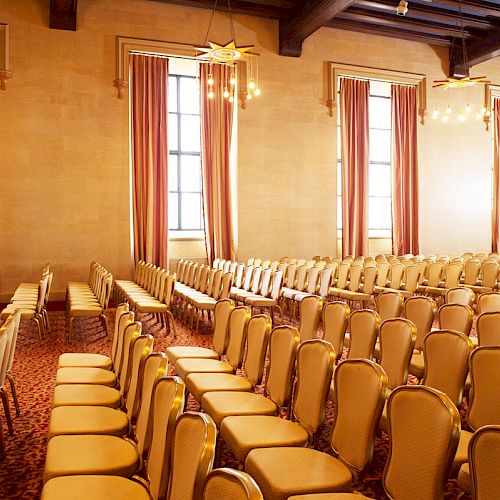 The image shows an empty room with rows of beige chairs, large windows with curtains, and chandeliers hanging from the ceiling.