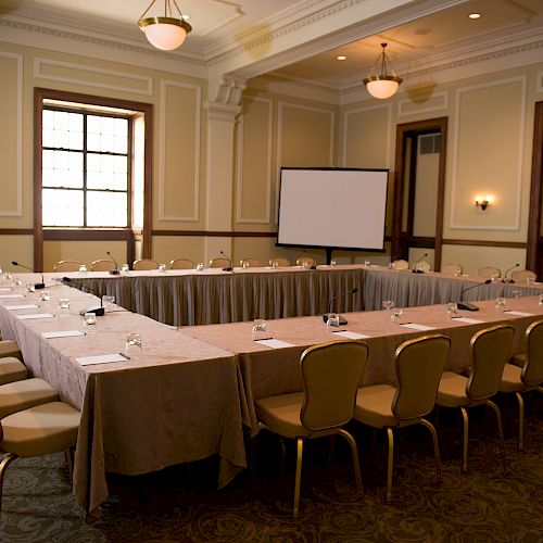 The image shows a conference room with a U-shaped table setup, chairs, a projector screen, and some notepads and pens on the tables.