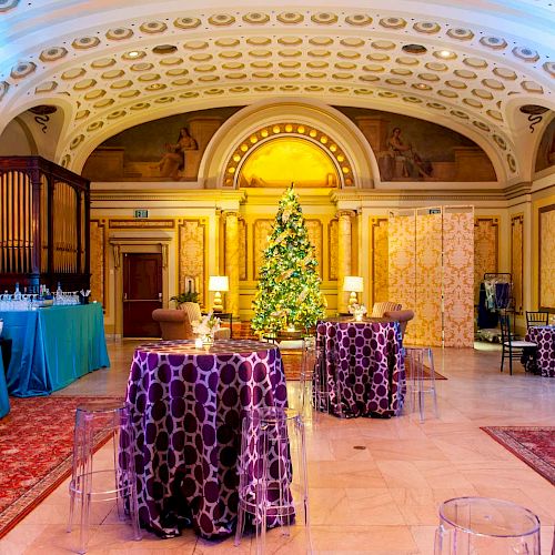 An elegantly decorated room with tables covered in purple cloths, a Christmas tree, food stations, ornate ceilings, and arched doorways.