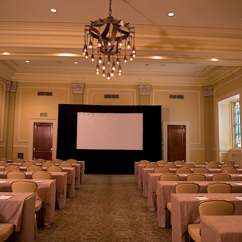 The image shows a well-lit, elegant conference room with rows of tables and chairs facing a large projection screen.