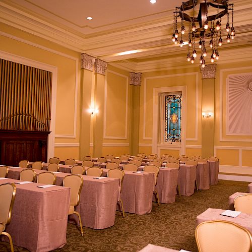 The image shows an elegant conference room with rows of tables and chairs, chandeliers, a stained glass window, and soft lighting.