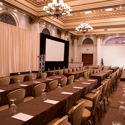 This image shows an elegant conference room with rows of tables and chairs, set up for a seminar or meeting, featuring chandeliers and decorated ceilings.