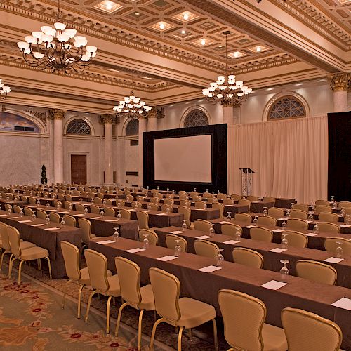 A large, elegant conference room with rows of tables and chairs facing two projection screens and a speaker's podium, adorned with chandeliers and ornate decor.