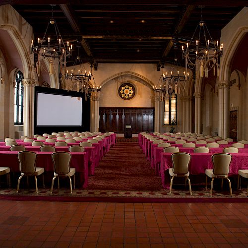 This image shows an ornate conference room set up with many chairs and tables facing a large projection screen at the front, under elegant chandeliers.