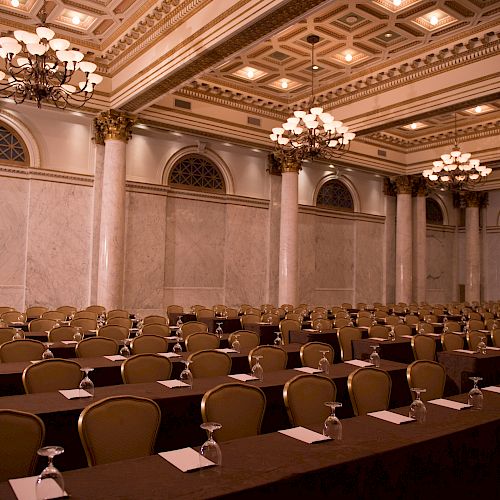 Elegant conference room with rows of tables and chairs, chandeliers, a high ceiling, and decorative columns. Photographer's watermark is on the corner.