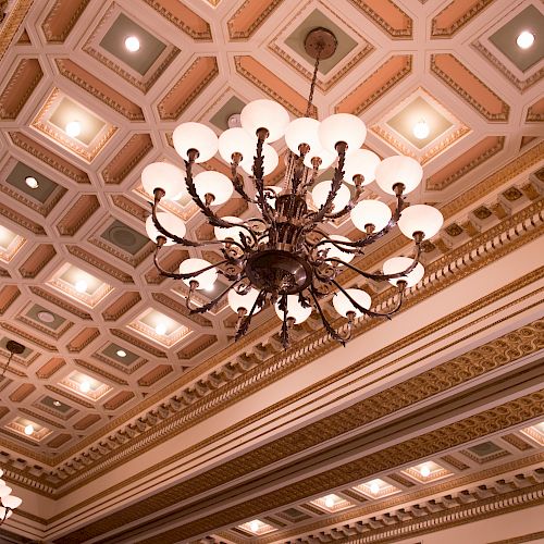 The image shows an ornate ceiling with intricate designs and several large chandeliers. The ceiling has recessed lighting.