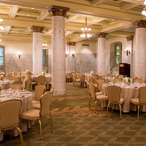 The image features an elegant banquet hall with round tables set for an event, decorated with floral centerpieces, chandeliers, and large marble columns.