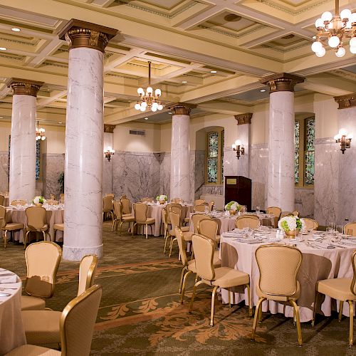 The image depicts an elegant banquet hall set up for an event with round tables, chairs, chandeliers, and marble columns.