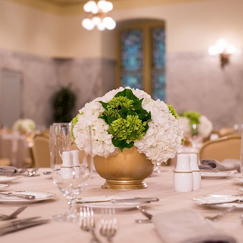 A formal dining setting with a centerpiece featuring white and green flowers in a gold vase, surrounded by neatly arranged glassware and utensils.