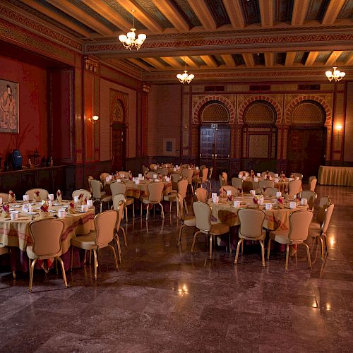 The image shows an elegant banquet hall with round tables set up with chairs and tableware. The hall has ornate decorations and chandeliers.