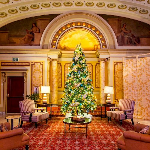 A lavishly decorated Christmas tree stands in a grand room with classical architecture, surrounded by armchairs and lamps on a red carpet.