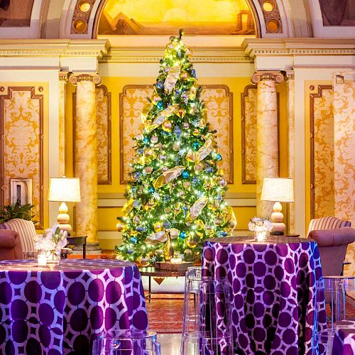 A decorated Christmas tree in an elegant interior with patterned tablecloths, chairs, and lamps on either side.