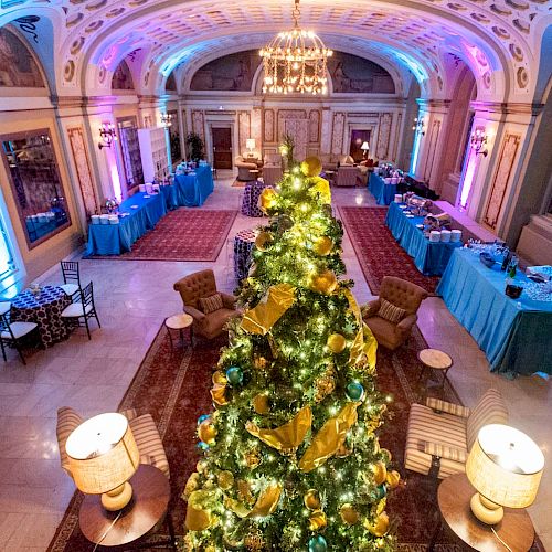 A grand hall with festive decor, featuring a large, decorated Christmas tree in the center, surrounded by tables and illuminated by chandeliers.