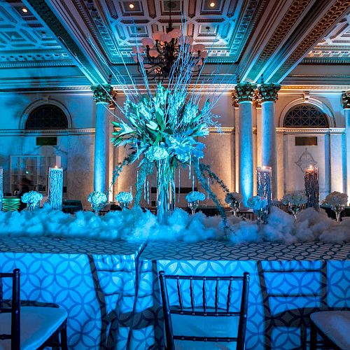 The image shows an elegantly decorated event hall with ornate columns, a large floral arrangement, and light blue lighting creating a sophisticated ambiance.