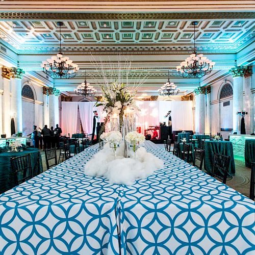 An elegant ballroom with ornate ceilings, chandeliers, and tables with blue patterned cloths. Floral centerpieces adorn the tables, and seating areas.