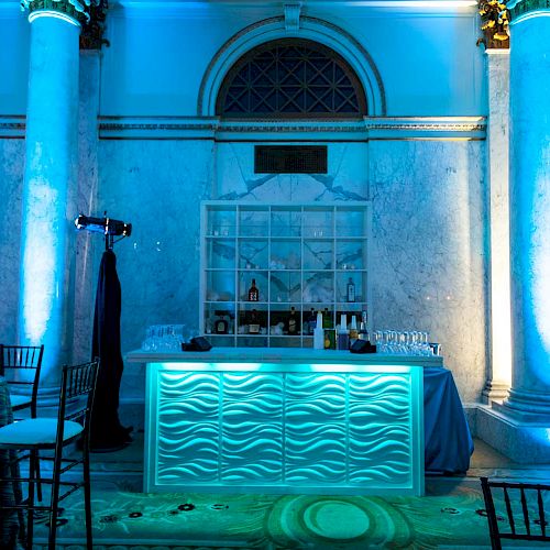 The image depicts a modern, illuminated bar set up in a grand hall with white marble columns, high ceilings, and blue lighting.