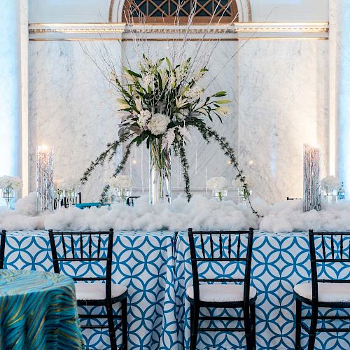 A beautifully decorated banquet table with a tall floral centerpiece, surrounded by black chairs, in a room with marble columns and blue accents.