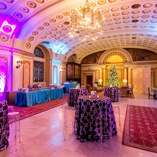 The image shows an elegantly decorated indoor venue with round tables, a grand piano, chandeliers, and a Christmas tree, featuring purple lighting.