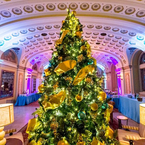 A decorated Christmas tree with green and gold ornaments stands in the center of an ornate room with arched ceilings and illuminated details.