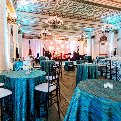 An elegant event setup in a grand hall with chandeliers, round tables with blue tablecloths, and chairs. The room is lit with warm and ambient lighting.