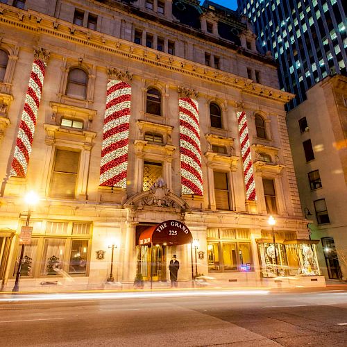 An ornate building with large, vertically striped red and white banners on its facade, streetlamps, and light trails from passing vehicles at dusk.