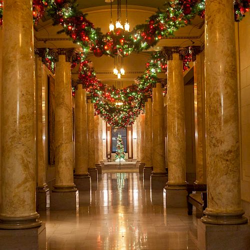 The image shows a decorated hallway with marble columns, lit chandeliers, and festive garlands hanging above, leading to a distant Christmas tree.