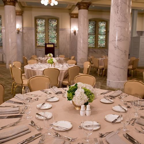 This image shows an elegant dining setup in a banquet hall with round tables, floral centerpieces, and beige chairs.