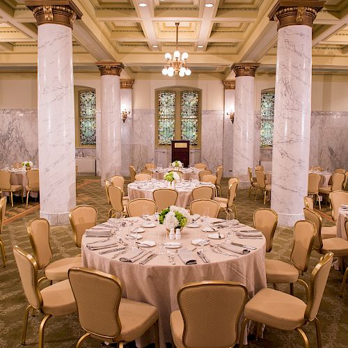 An elegantly decorated banquet hall with round tables, beige chairs, floral centerpieces, marble columns, chandeliers, and carpeted floor.