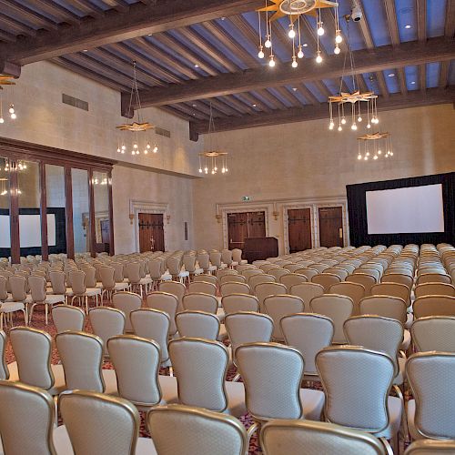 An empty conference room with rows of beige chairs, chandeliers on the ceiling, and a large projection screen at the front.