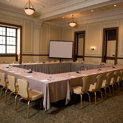 An empty conference room with U-shaped table setup, chairs, tablecloths, water glasses, and a projection screen. The room is well-lit with elegant decor.