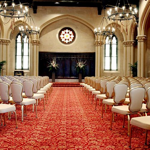 An elegant hall with rows of white chairs, red carpet, chandeliers, arched windows, and a stage adorned with floral arrangements, set for an event.