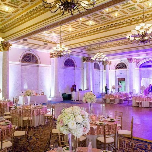 This image shows an elegantly decorated ballroom with round tables, floral centerpieces, chandeliers, and a stage with purple lighting, ready for an event.