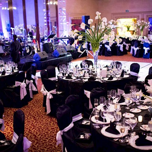 This image shows a decorated banquet hall with round tables, chairs with black covers and white bows, glassware, and floral centerpieces.