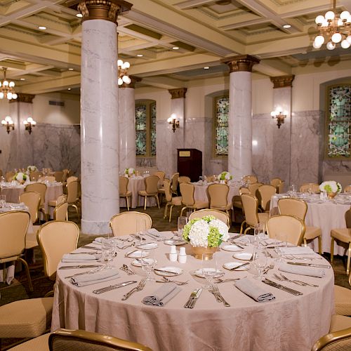 A beautifully decorated ballroom with round tables set for an event, featuring elegant chairs, floral centerpieces, and intricate ceiling details.