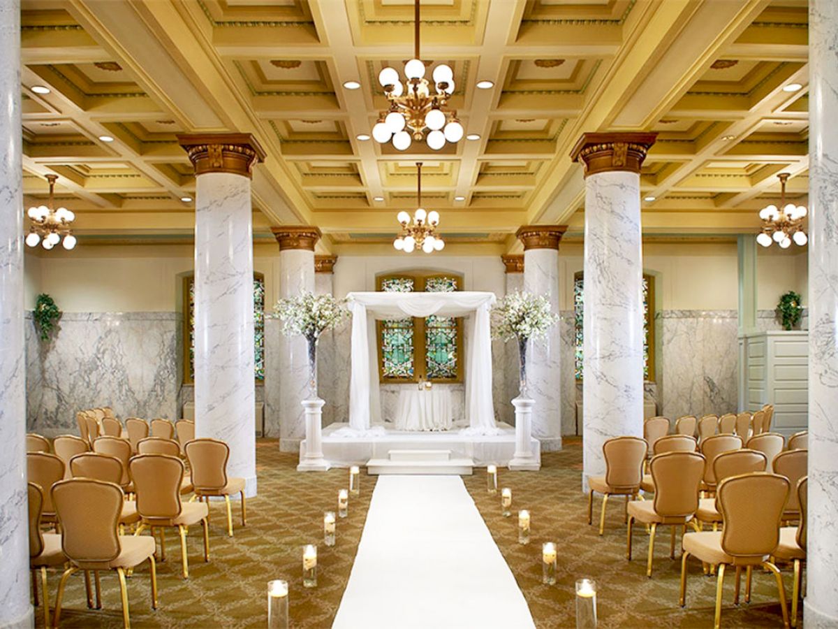 This image shows an elegant indoor wedding ceremony setup with marble columns, golden chairs, a white aisle, floral decorations, and chandeliers.